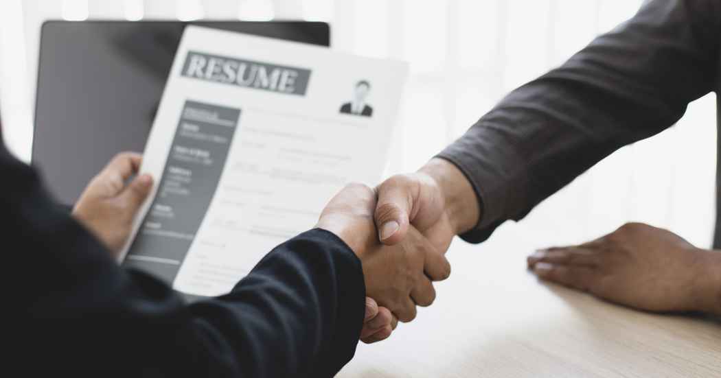 Two people shake hands over a resume on a desk, signaling a job offer or agreement during a meeting with a recruiter.