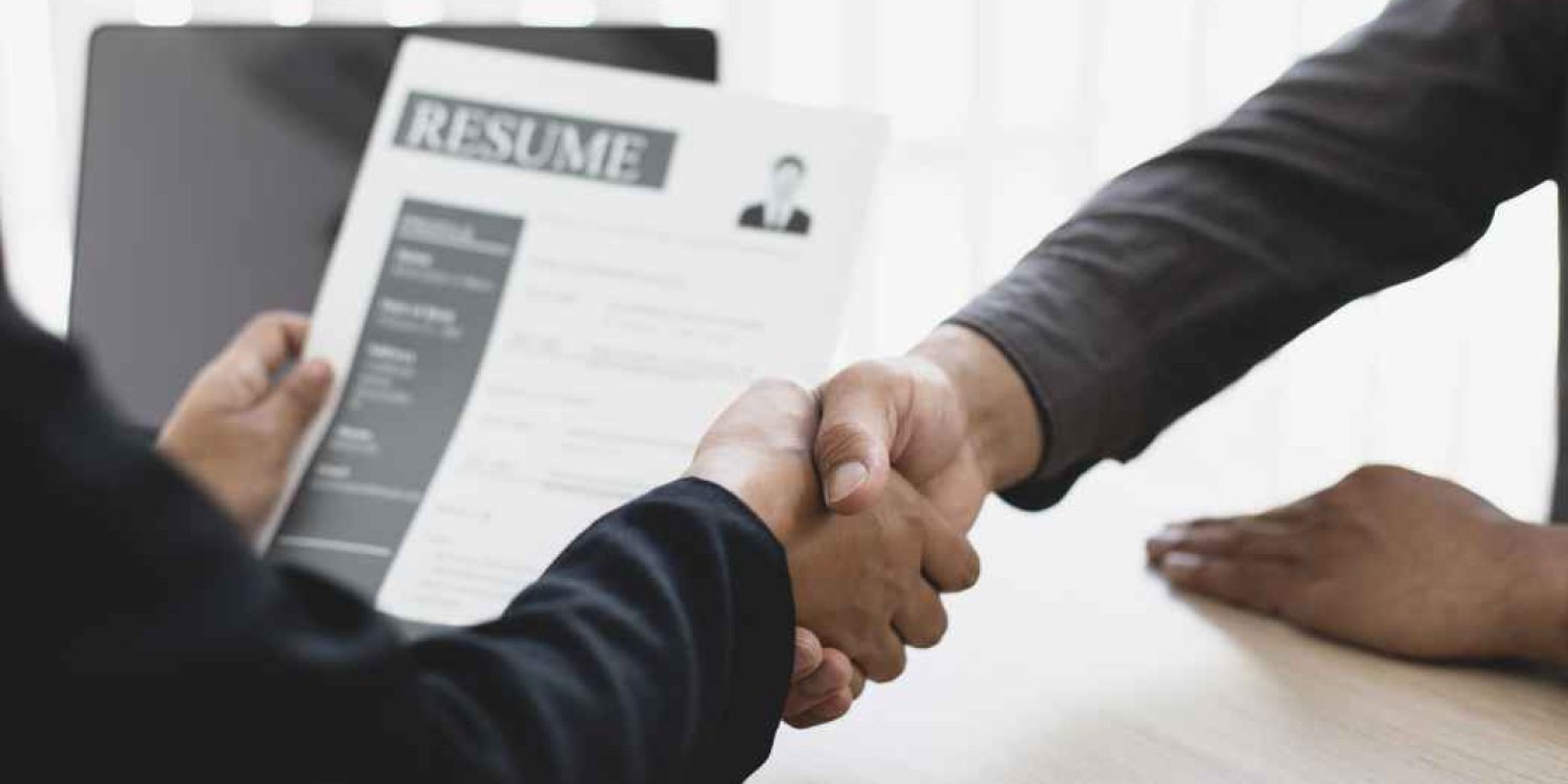 Two people shake hands over a resume on a desk, signaling a job offer or agreement during a meeting with a recruiter.